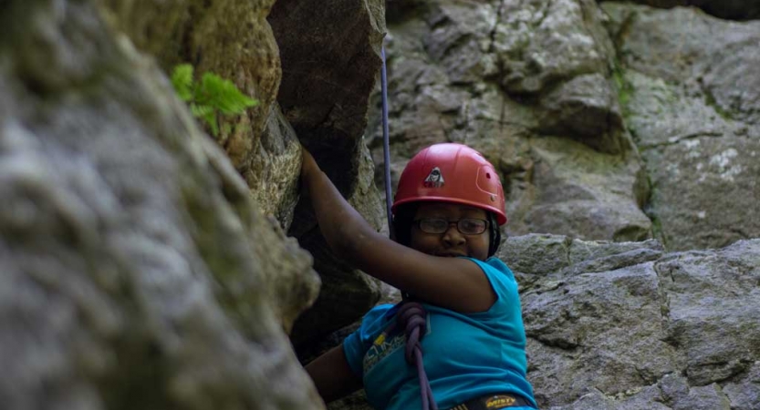 A person wearing safety gear is secured by ropes as they look down at the camera and smile while rock climbing.